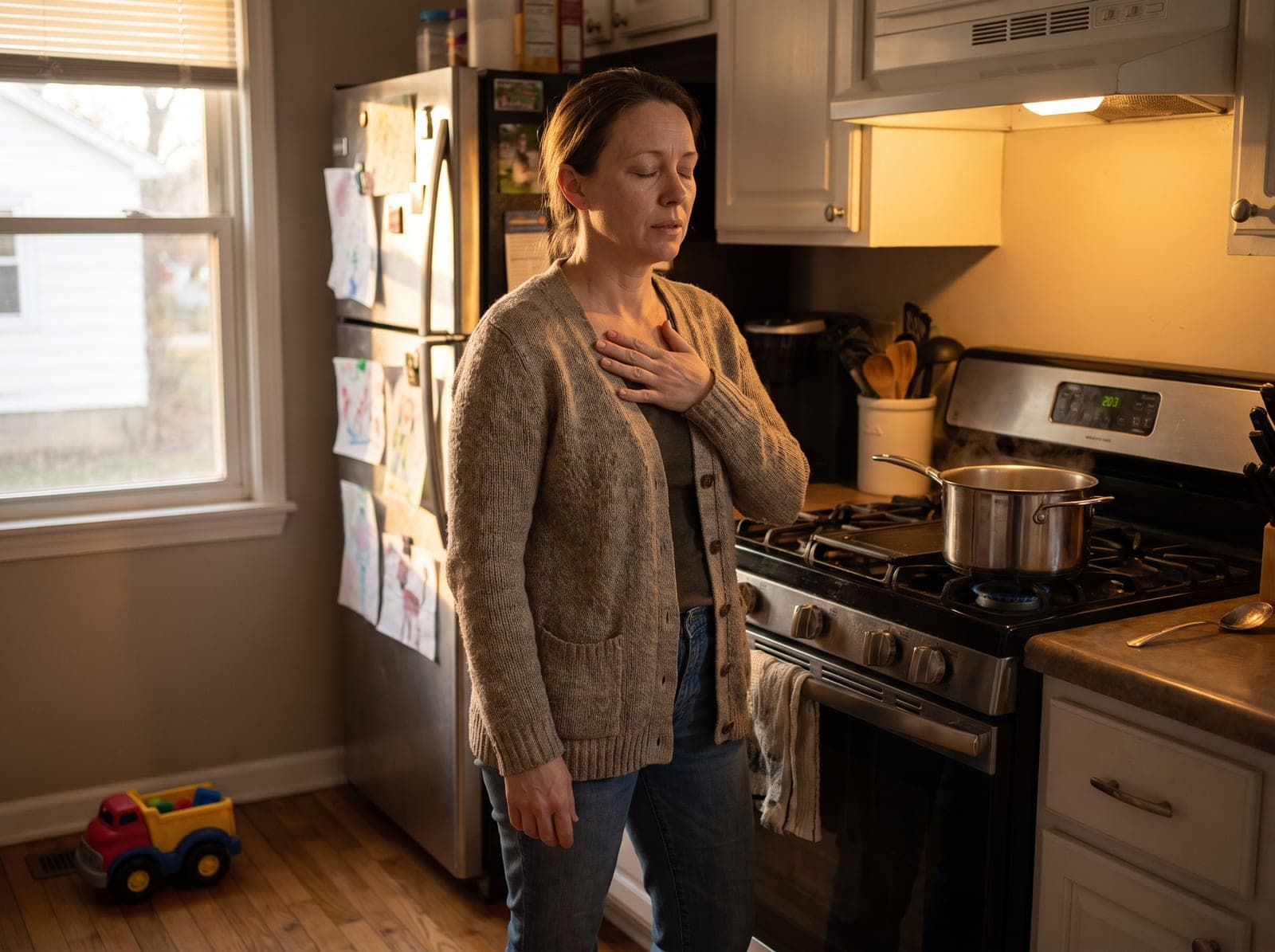 A mother stands alone at her kitchen counter at dusk, one hand on her chest, eyes closed, breathing out.